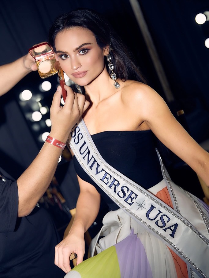 A Miss USA contestant has her makeup finished backstage under the lights as a compact and lip color are held to her face.