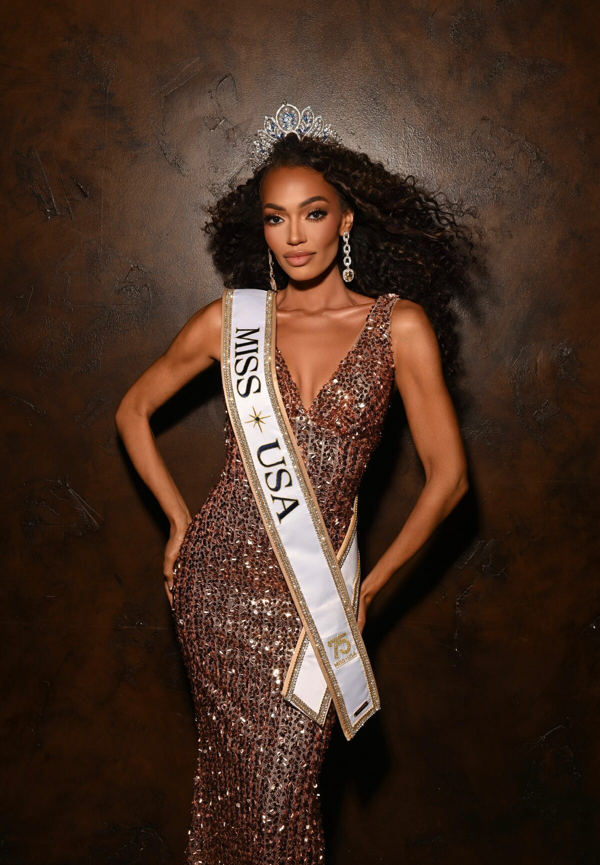 Miss USA Justus Kelley in a bronze sequin gown, crown, and crystal drop earrings against a dark backdrop.