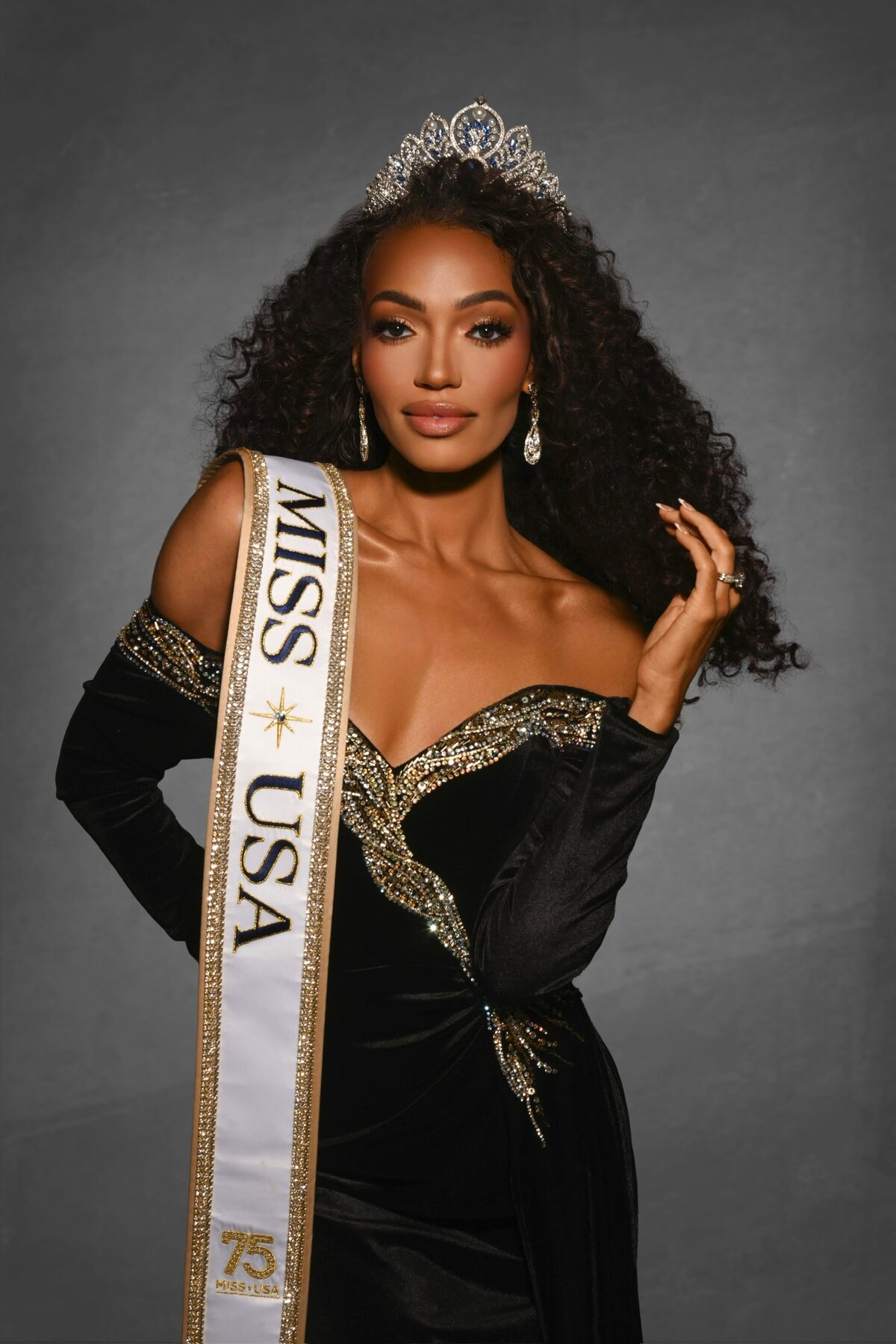 Miss USA Justus Kelley in a black velvet gown, crown, and long crystal drop earrings against a soft grey backdrop.