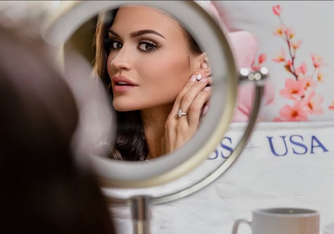 A contestant fastens a teardrop earring, reflected in a round vanity mirror beside a Miss USA banner and a spray of blossoms.